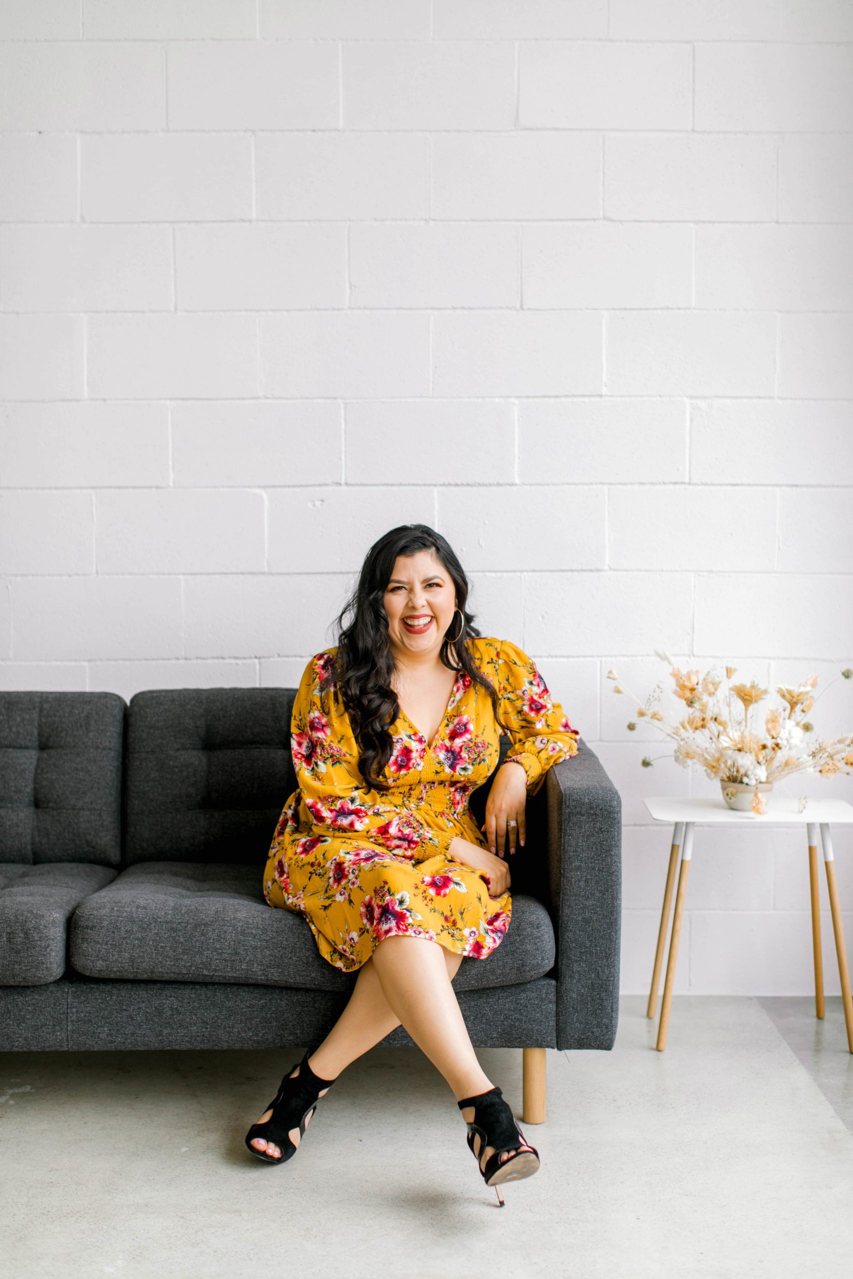 Full body picture of Latina laughing on a gray couch beside a dried flower arrangement on top of small nordic table
